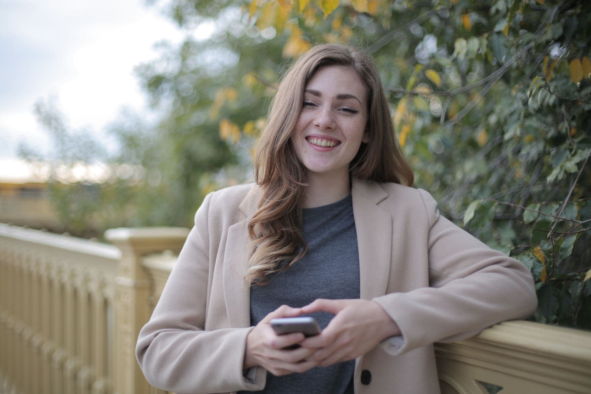 Uma mulher sorri com um celular nas mãos, apoiada em um corrimão ao ar livre.