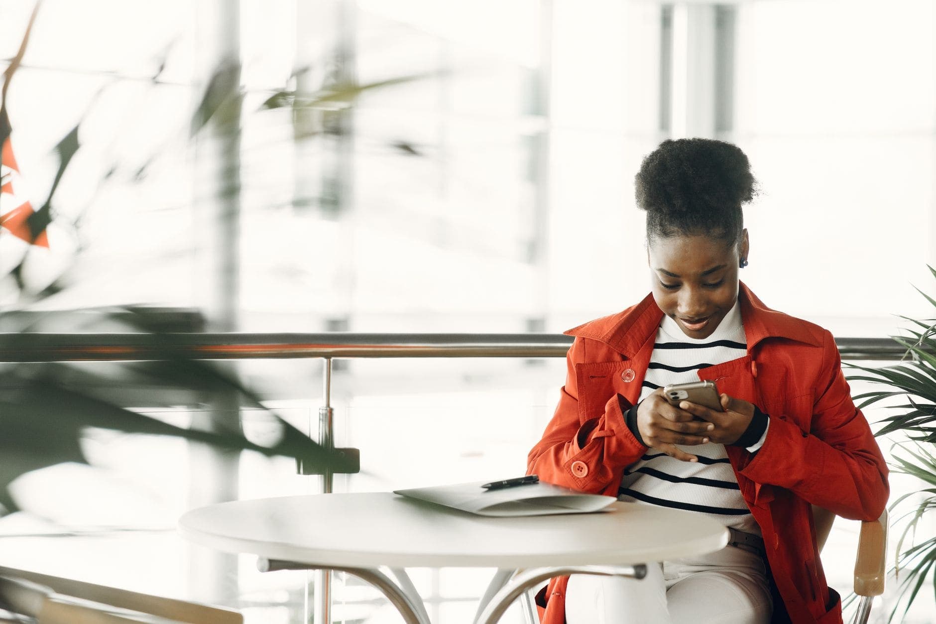 A woman in an orange coat looks at her phone at a round table beside a bright window.