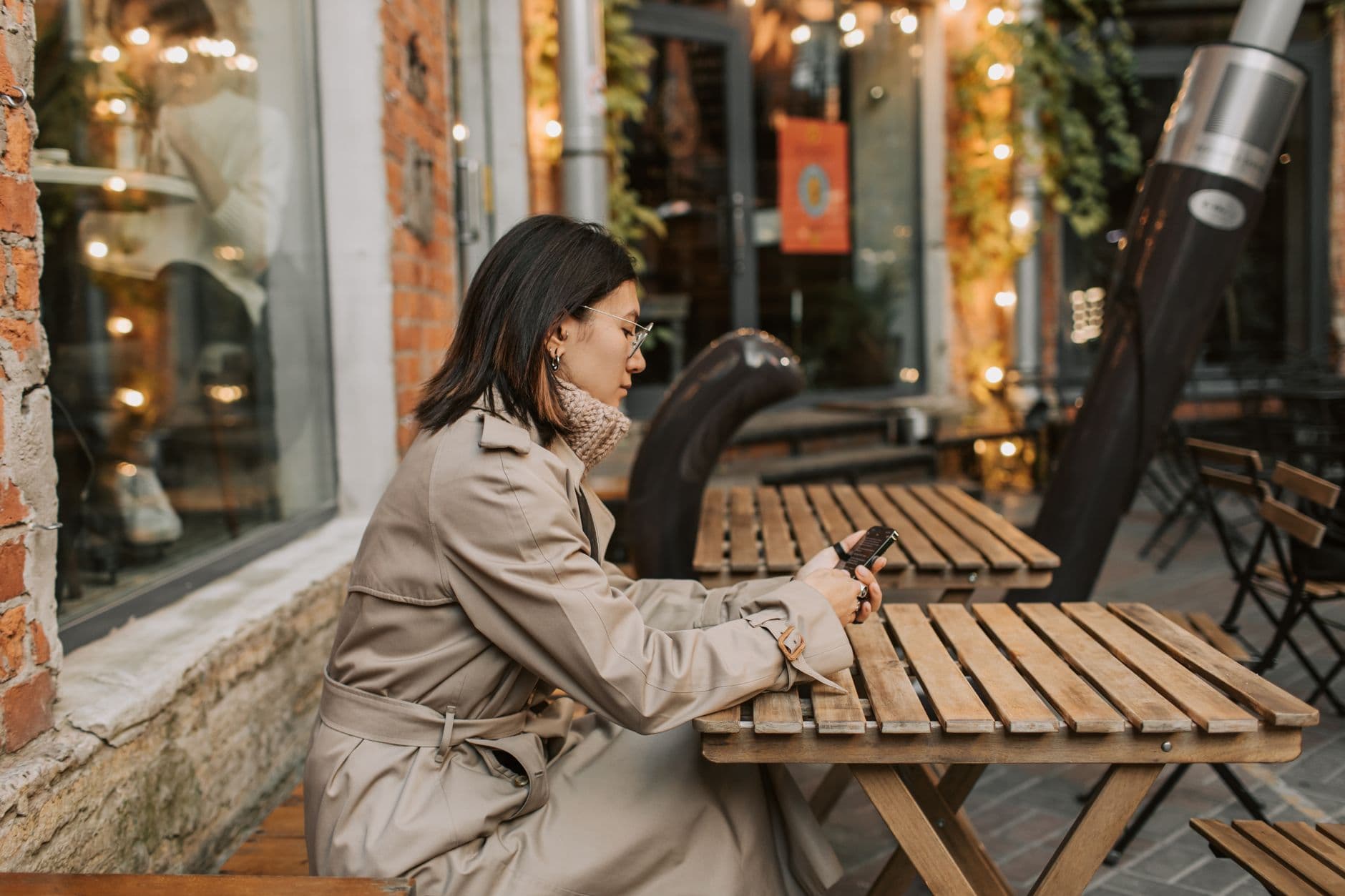 Eine Frau mit Brille tippt am Smartphone, an einem Tisch vor einem Café.