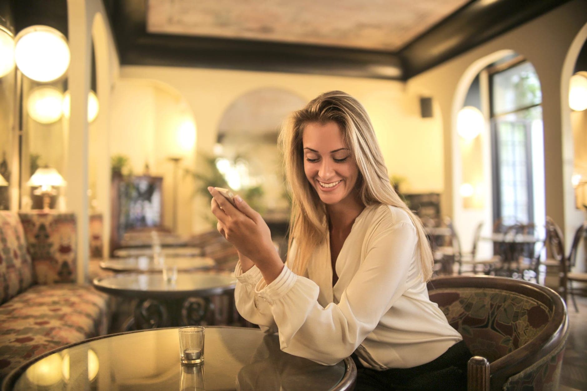 Moteris šviesia palaidine šypsodamasi žiūri į telefoną prie apvalaus kavinės stalo.