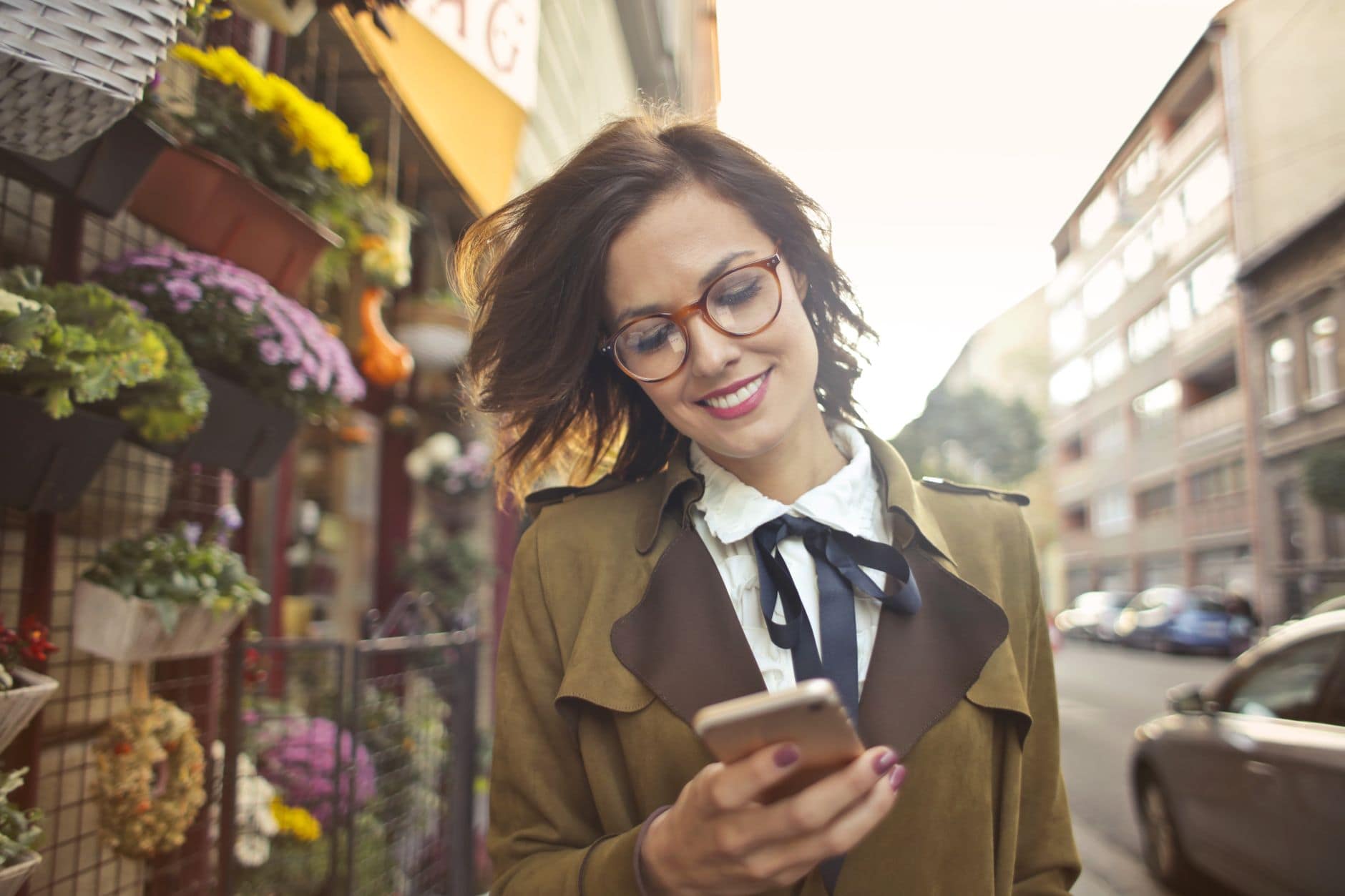 Eine lächelnde Frau mit Brille schaut vor einem Blumenladen in der Stadt auf ihr Smartphone.