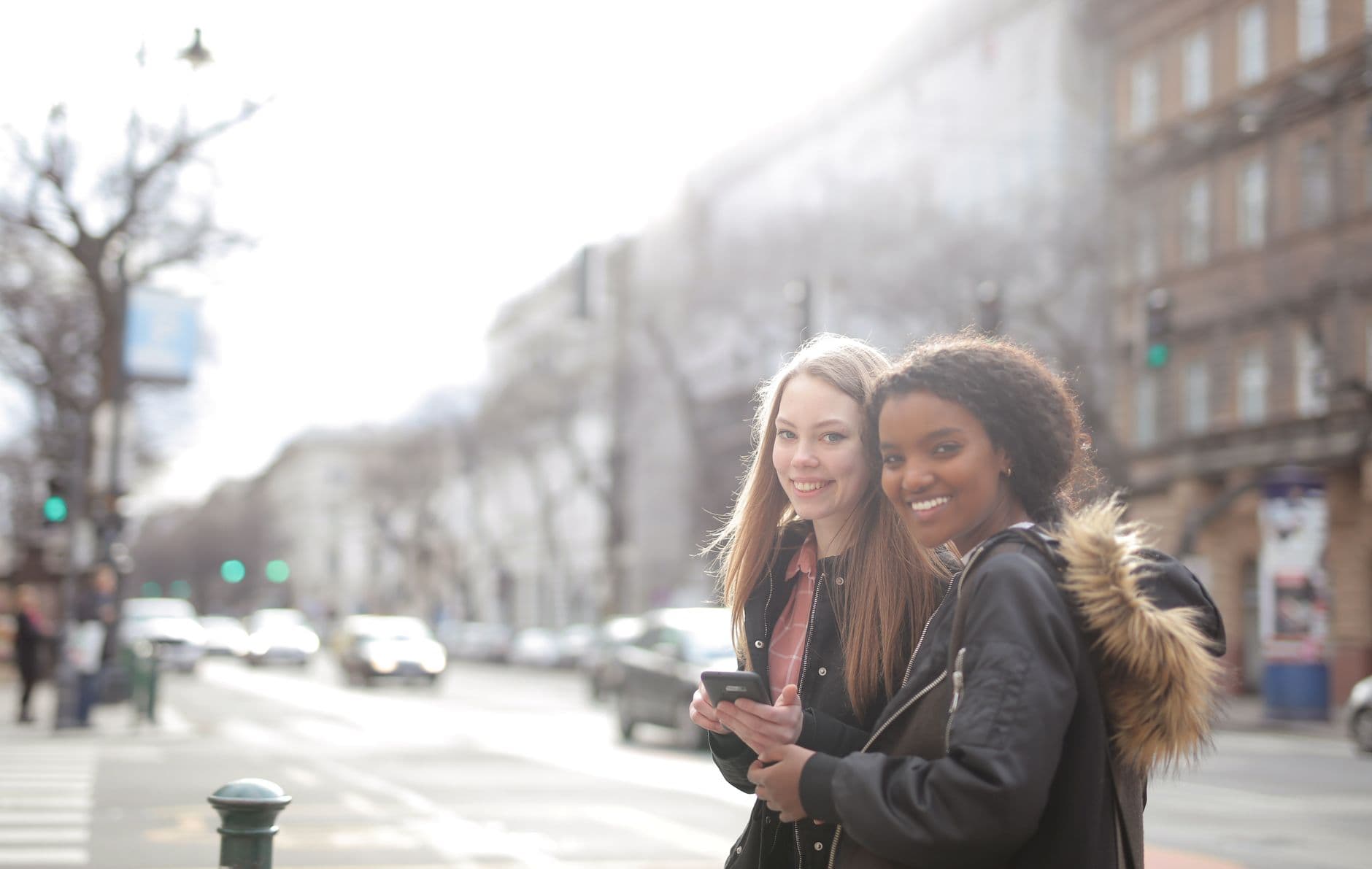 Deux jeunes femmes sourient dans une rue animée, l'une tenant un téléphone à la main.
