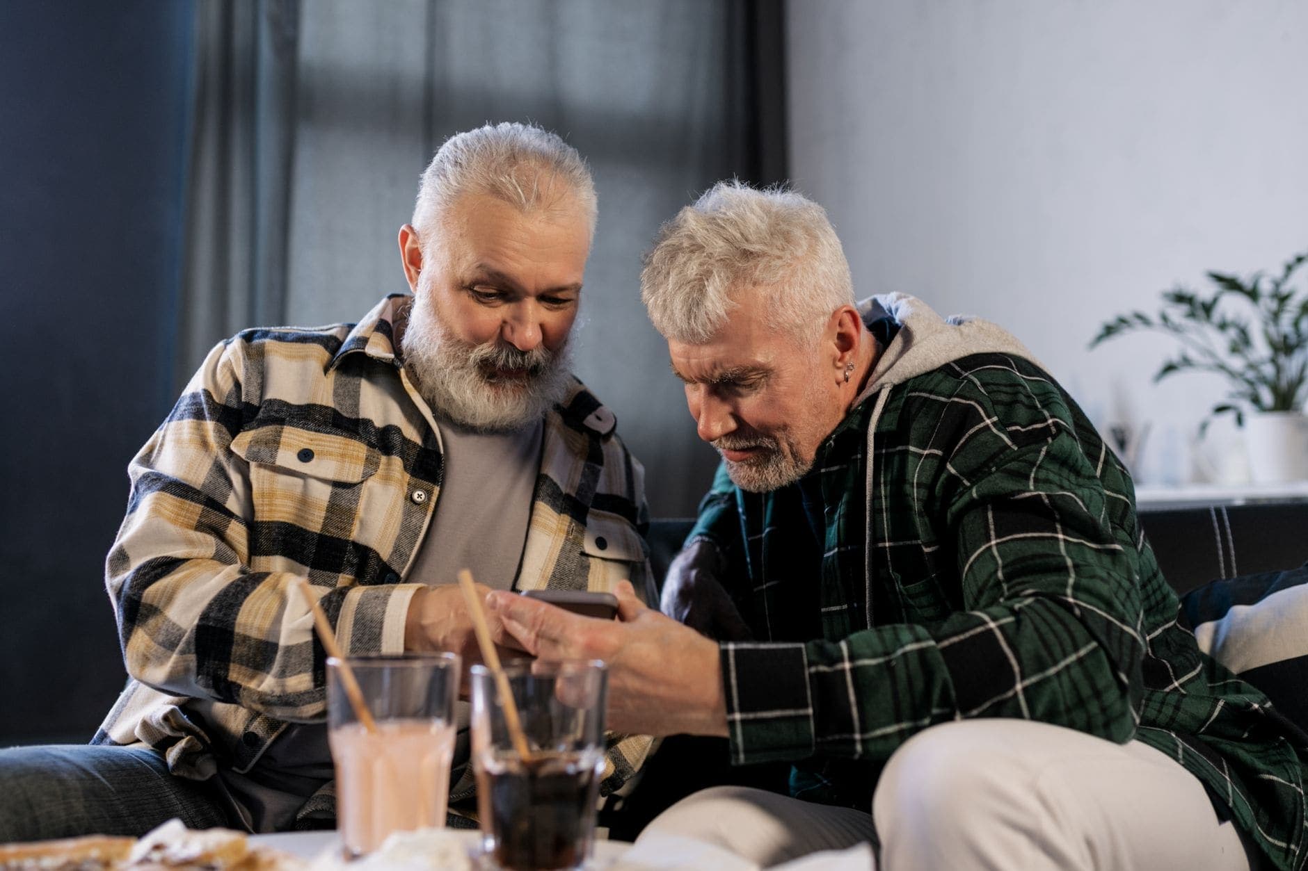 Two men in checked shirts look at a phone together, with two drinks on the table.