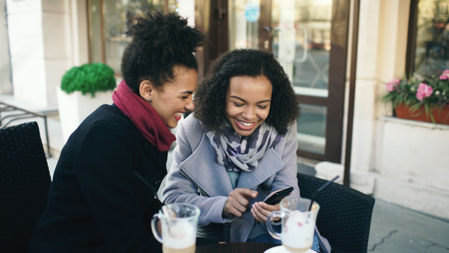 Due giovani donne ridono a un tavolino all'aperto guardando insieme uno smartphone.