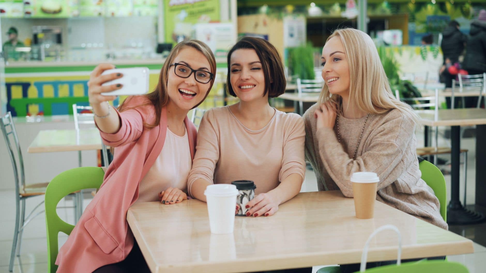 Trzy uśmiechnięte kobiety robią wspólne selfie przy stoliku z kubkami kawy