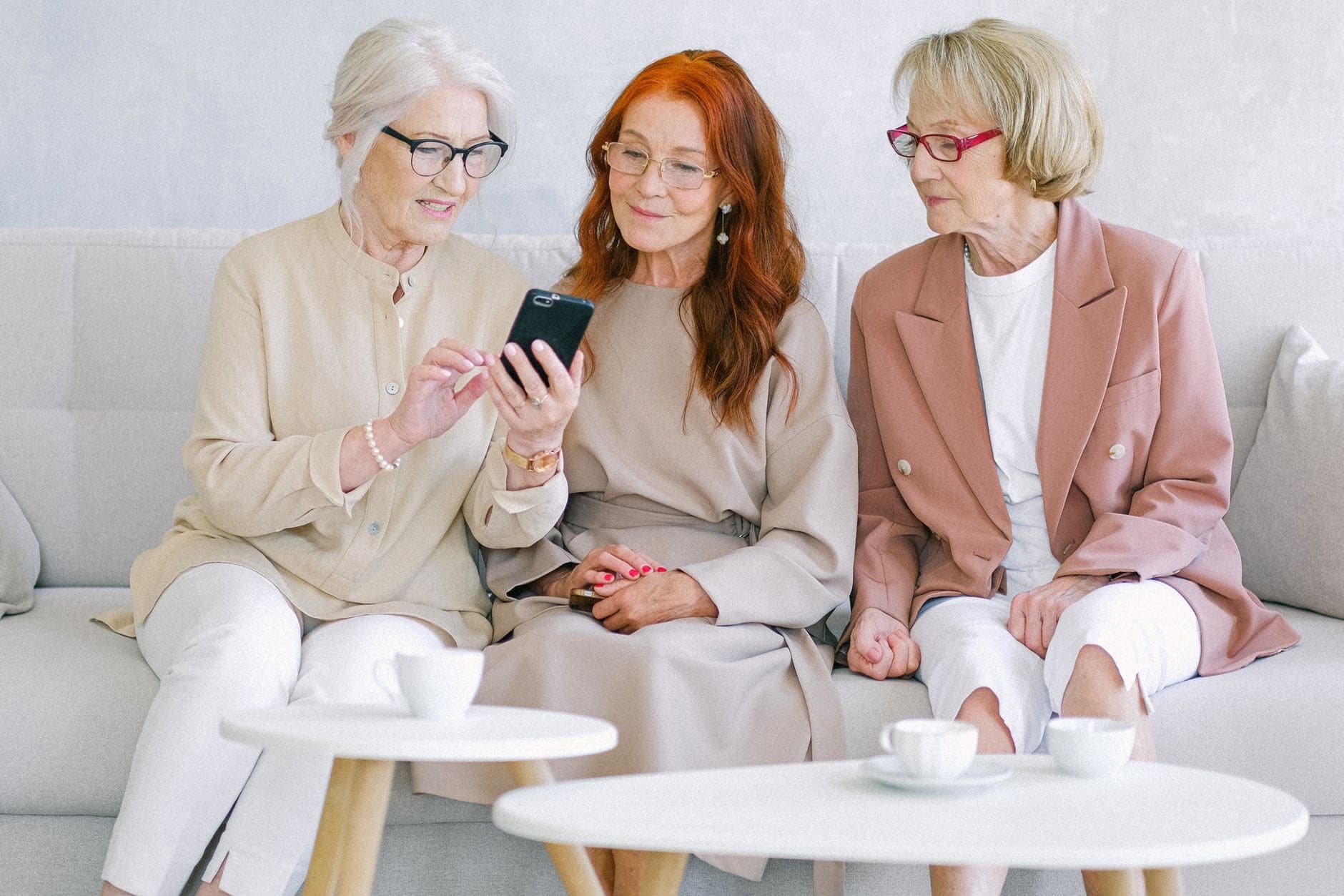 Três mulheres sentadas num sofá olham para um telemóvel junto a mesas com chávenas.