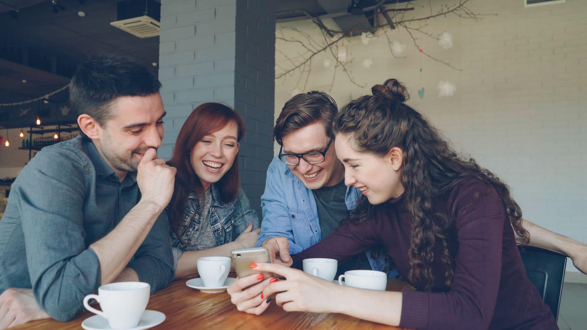 Vier Erwachsene lachen an einem Cafétisch und schauen gemeinsam auf ein Smartphone.