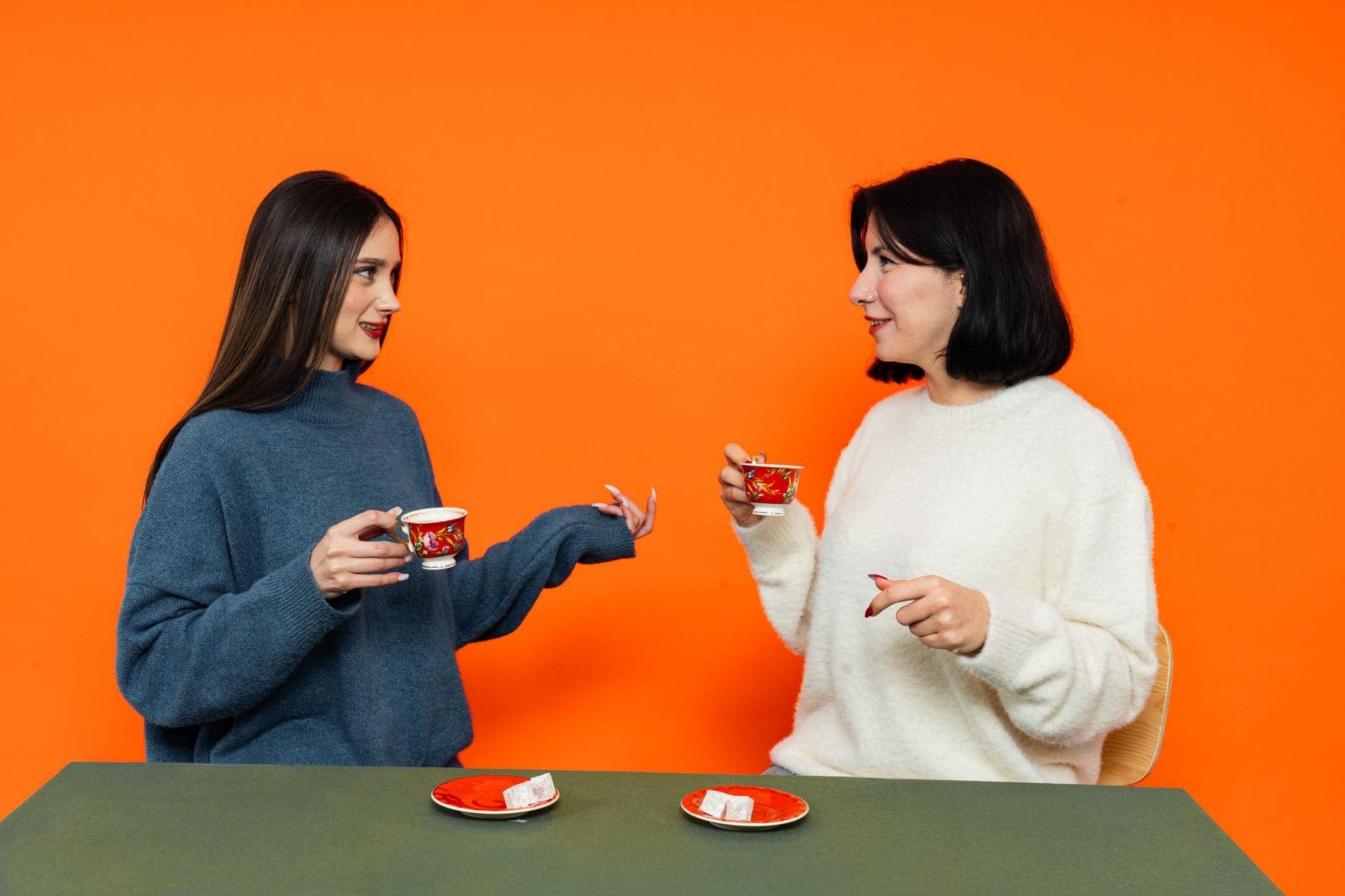 Two women in blue and cream jumpers hold red cups as they face each other against an orange backdrop.