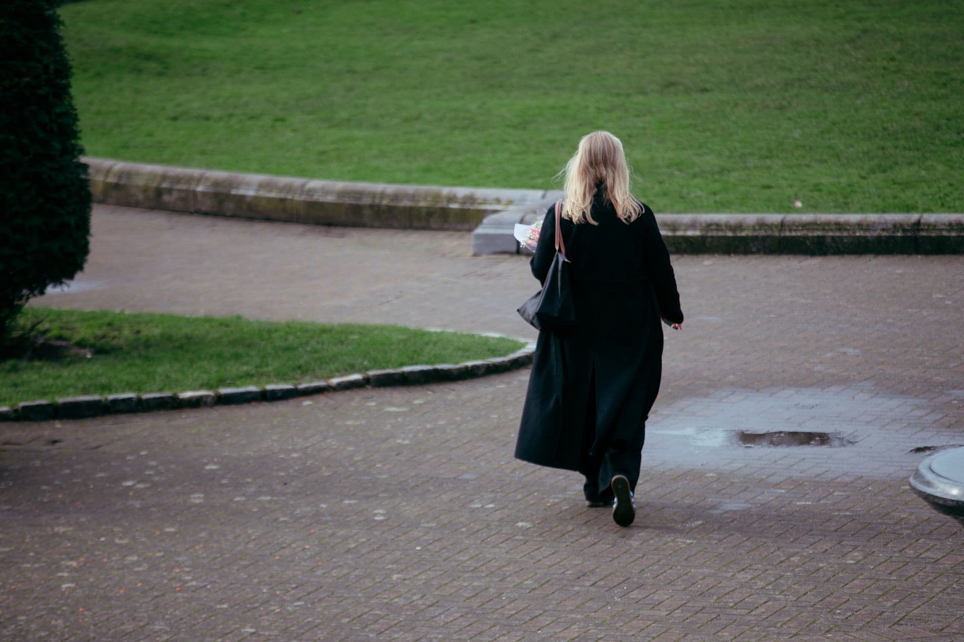 Une femme en manteau noir s'éloigne sur un chemin de parc, vue de dos, un bouquet à la main.