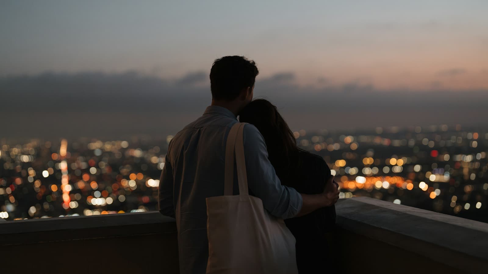 Couple vu de dos, enlacé au bord d'une terrasse, face aux lumières d'une ville au crépuscule