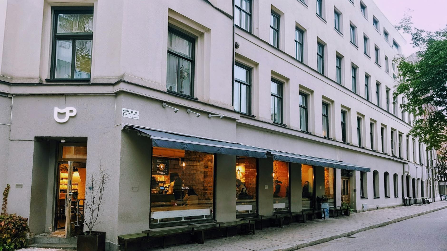 Hörnkafé med stora fönster och varmt ljus inne, i en vit huslänga längs en gata.