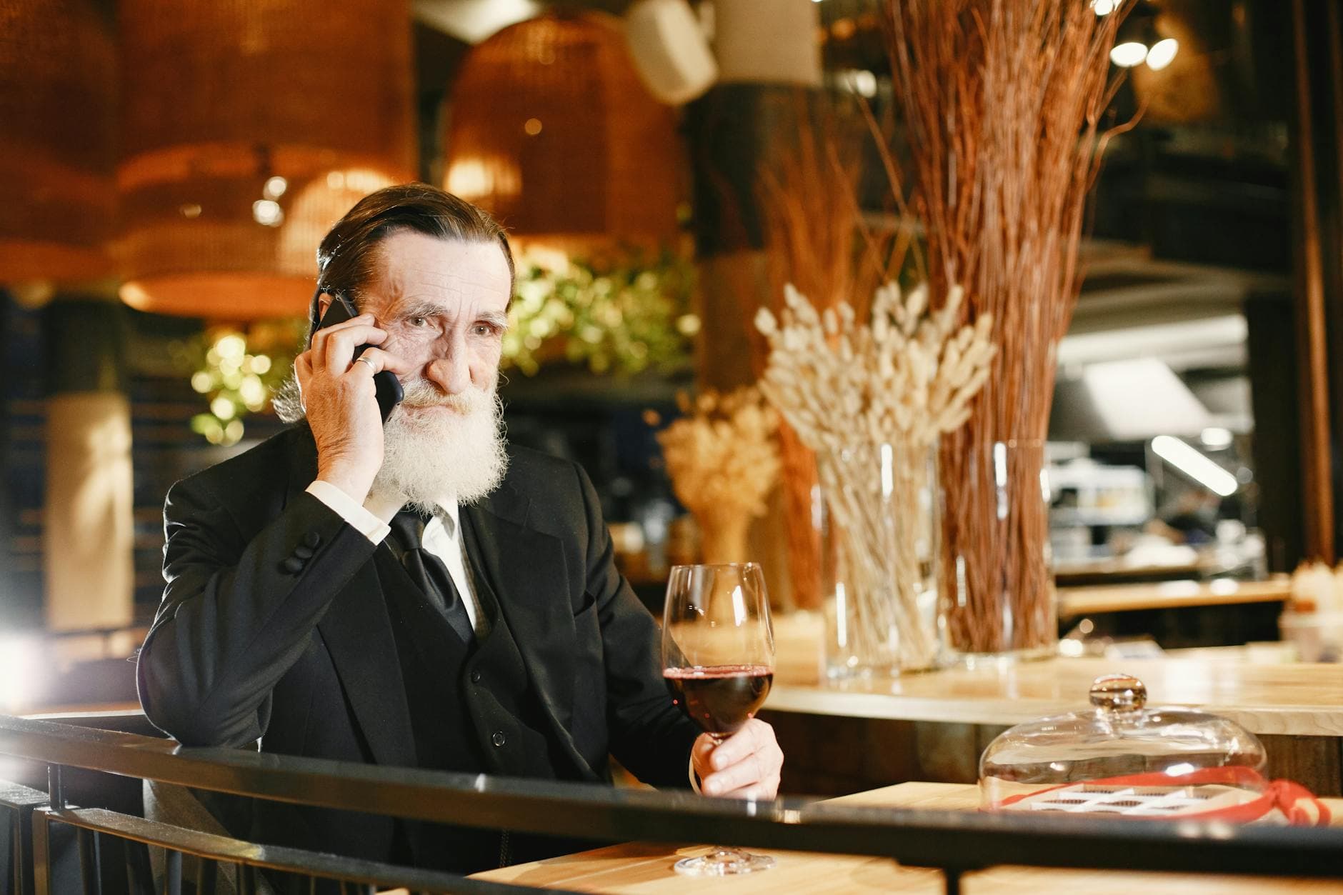 Man med vitt skägg i svart kostym talar i mobilen med ett glas rödvin på restaurang.