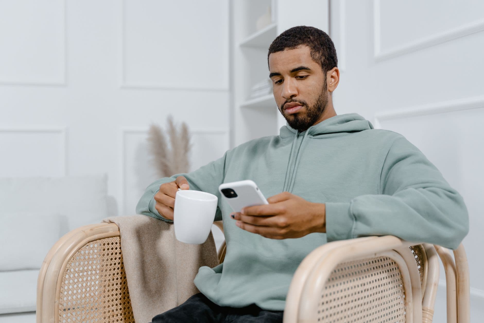 Un homme en sweat vert clair tient une tasse et un téléphone, assis dans un fauteuil.
