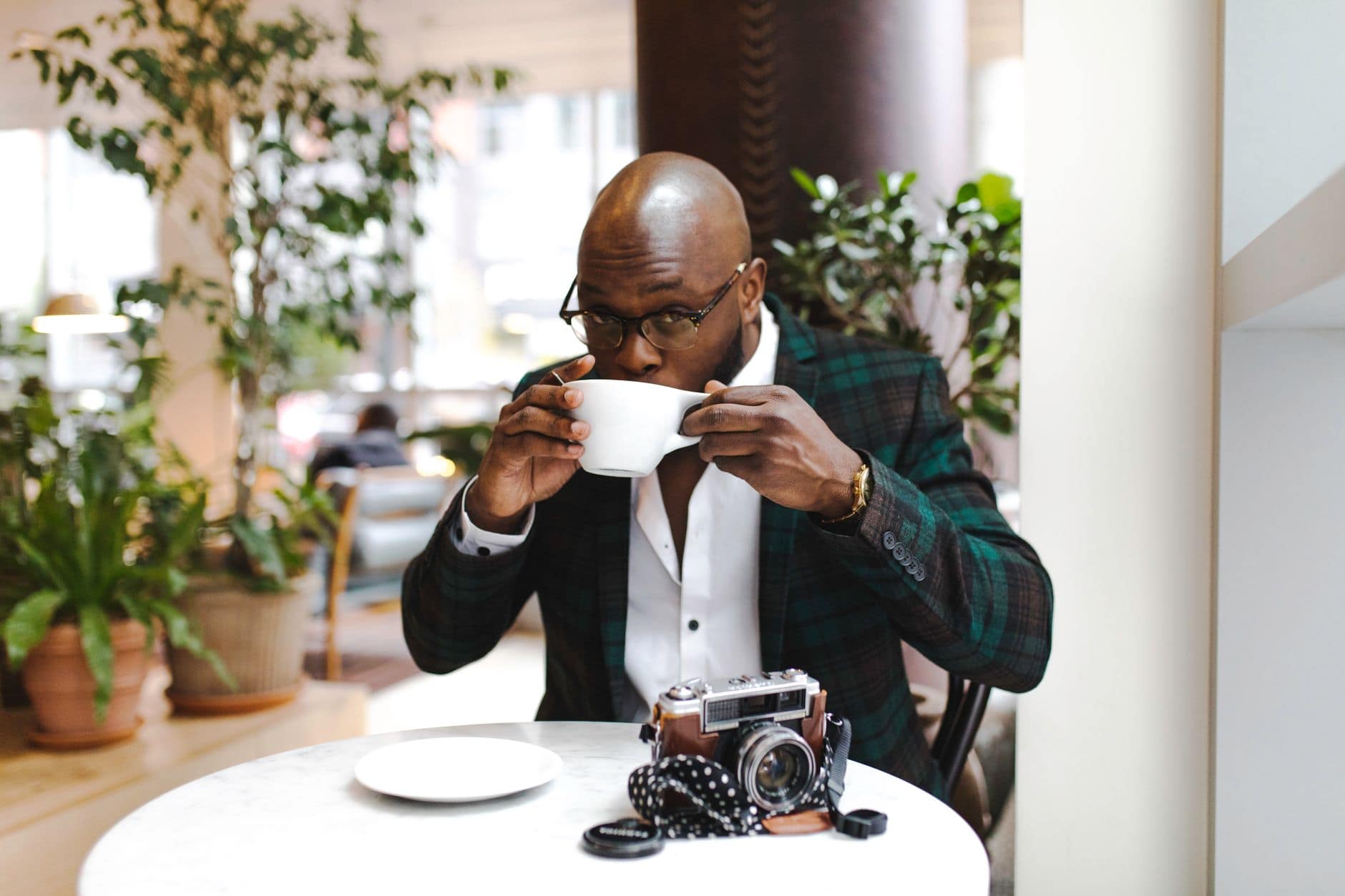 Un homme en veste à carreaux boit dans une tasse blanche, avec un appareil photo sur la table.