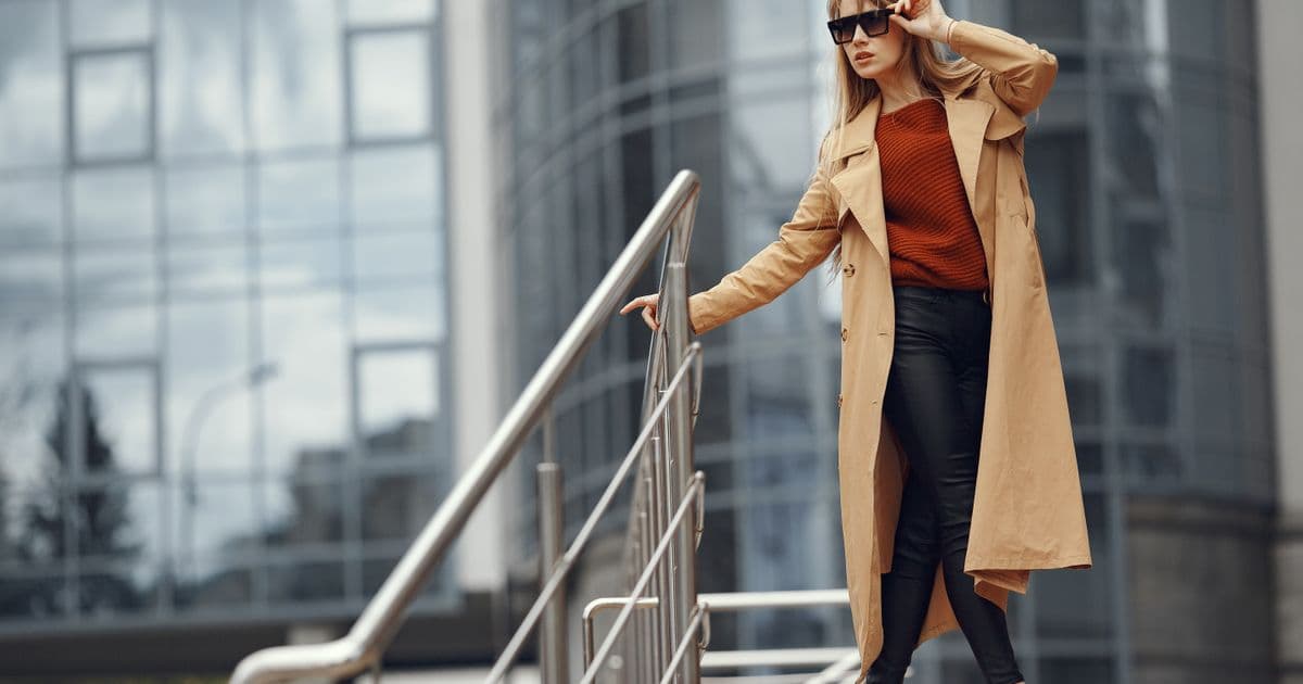 Frau mit Sonnenbrille und beigem Trenchcoat steht an einem Treppengeländer vor einer Glasfassade.