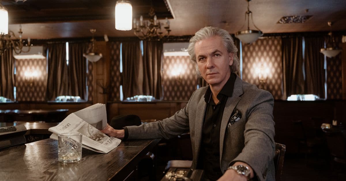 Mann mit grauen Haaren und grauem Sakko sitzt mit Zeitung und Glas an einer holzvertäfelten Bar.