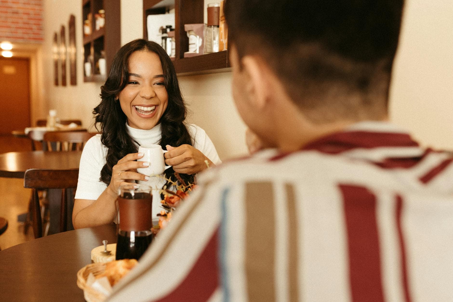 Vrouw met donker golvend haar lacht met een kopje in haar handen tegenover iemand in een café.
