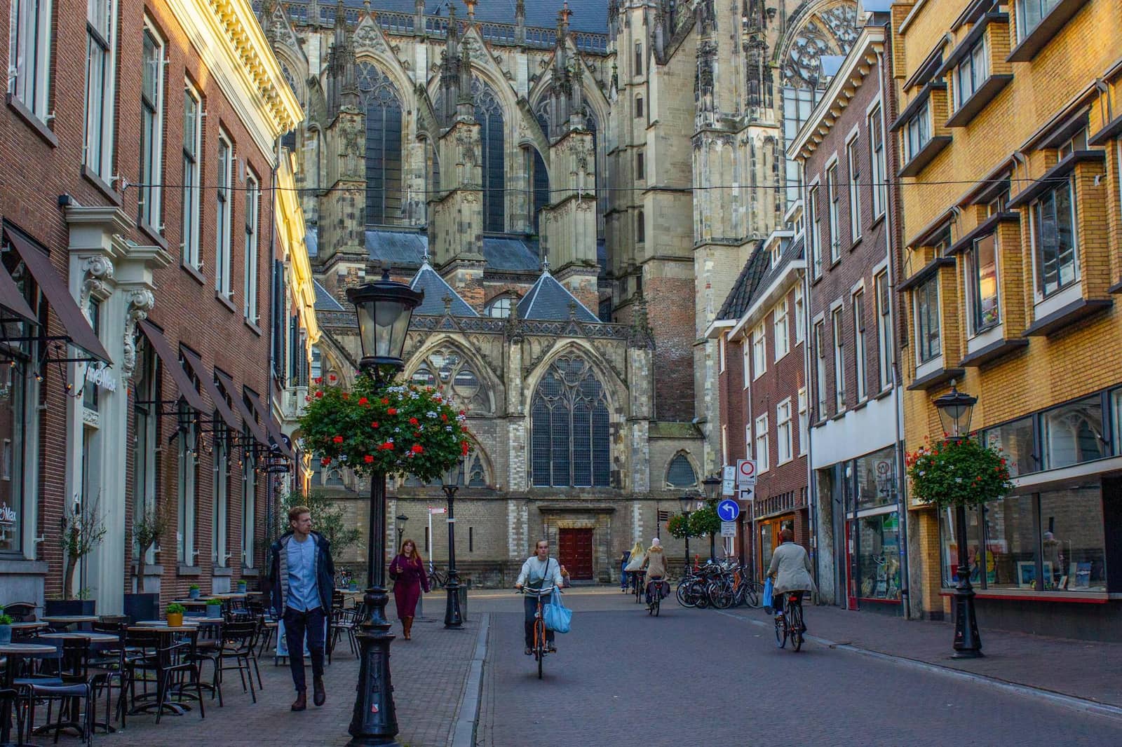Fietsers op een geplaveide straat voor een grote gotische kerk met bloembakken aan lantaarns.