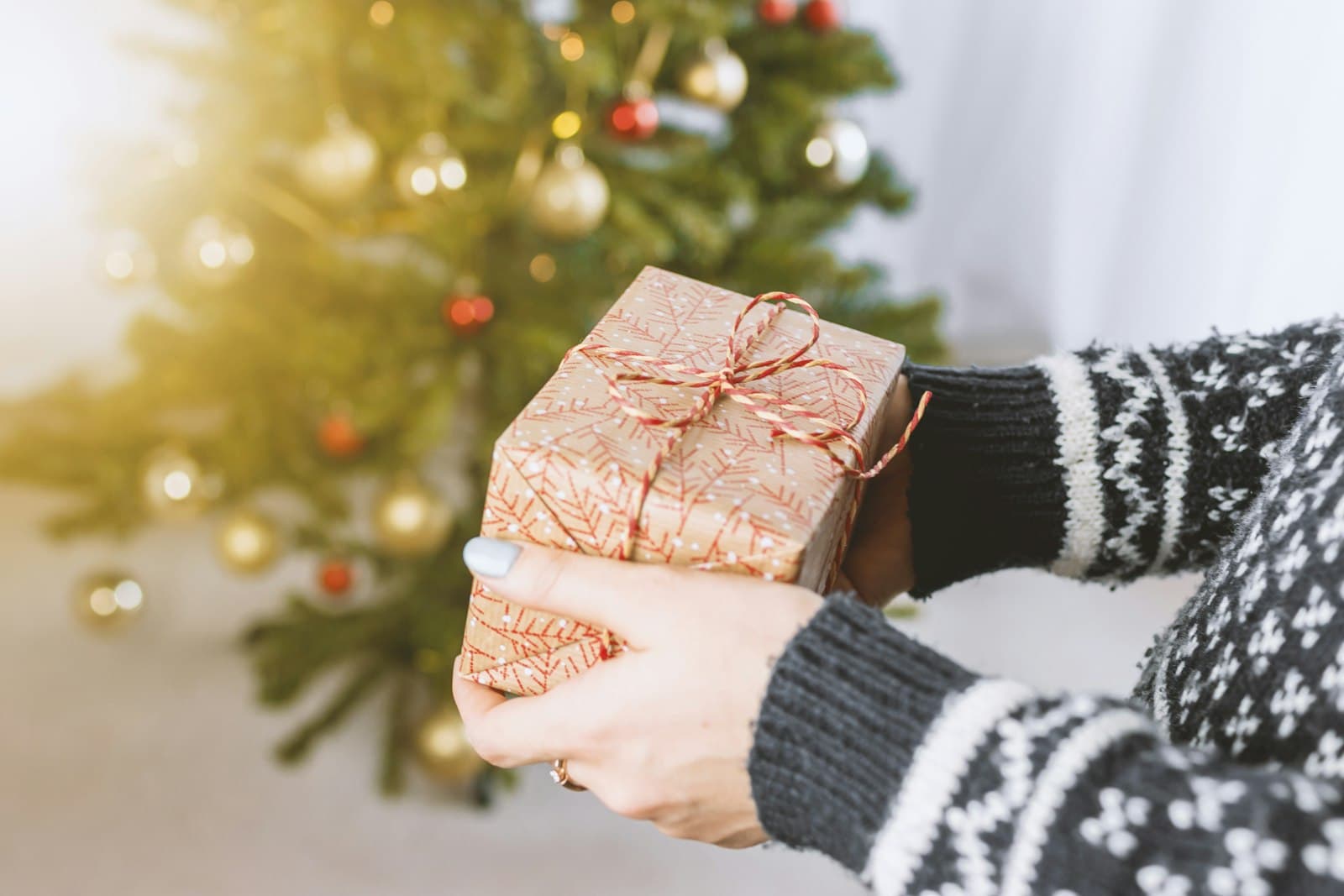 Hände im Norwegerpullover halten ein verpacktes Geschenk vor einem Weihnachtsbaum