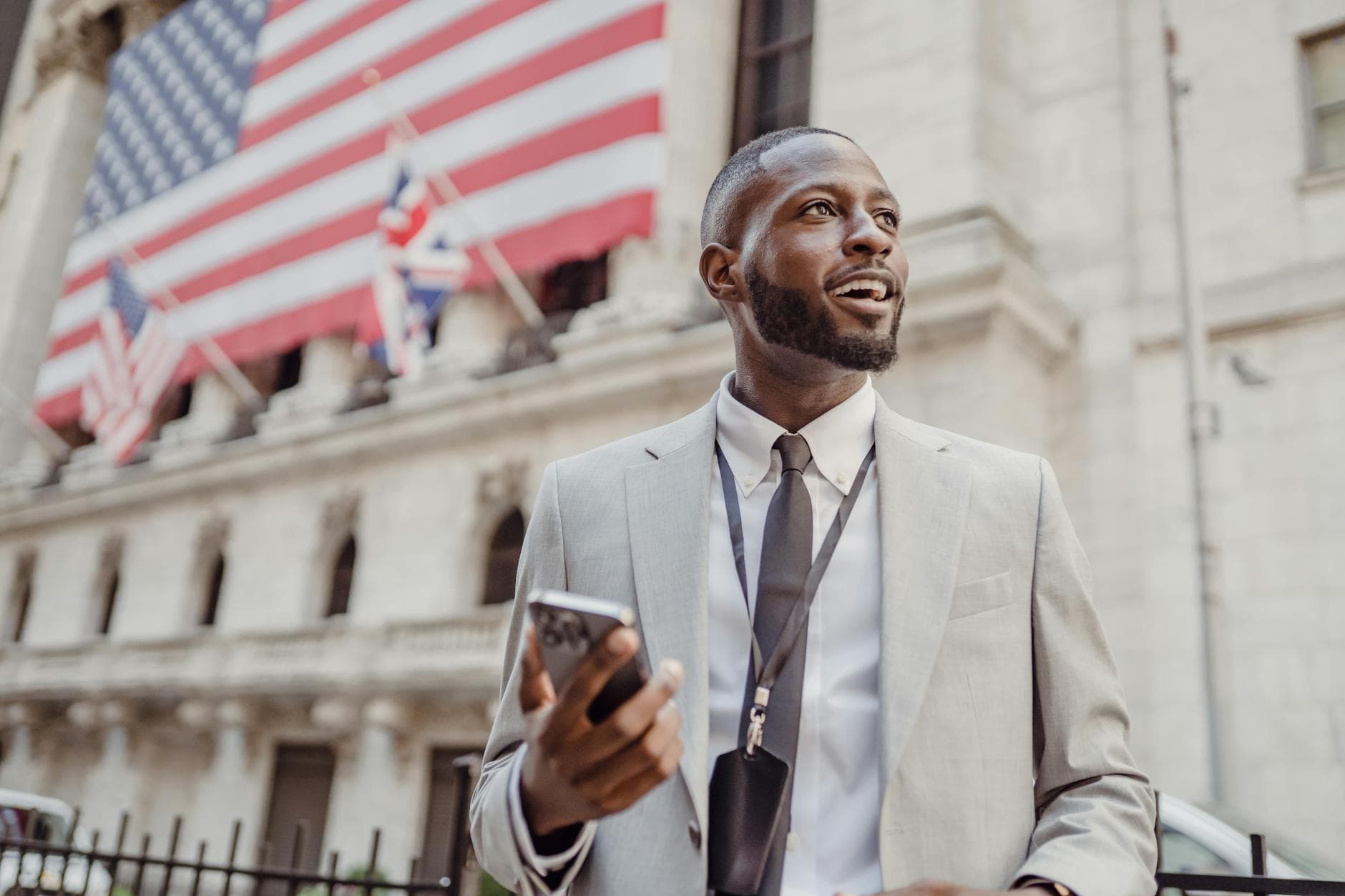 A man in a light gray suit holds a phone outside a stone building hung with a large flag.