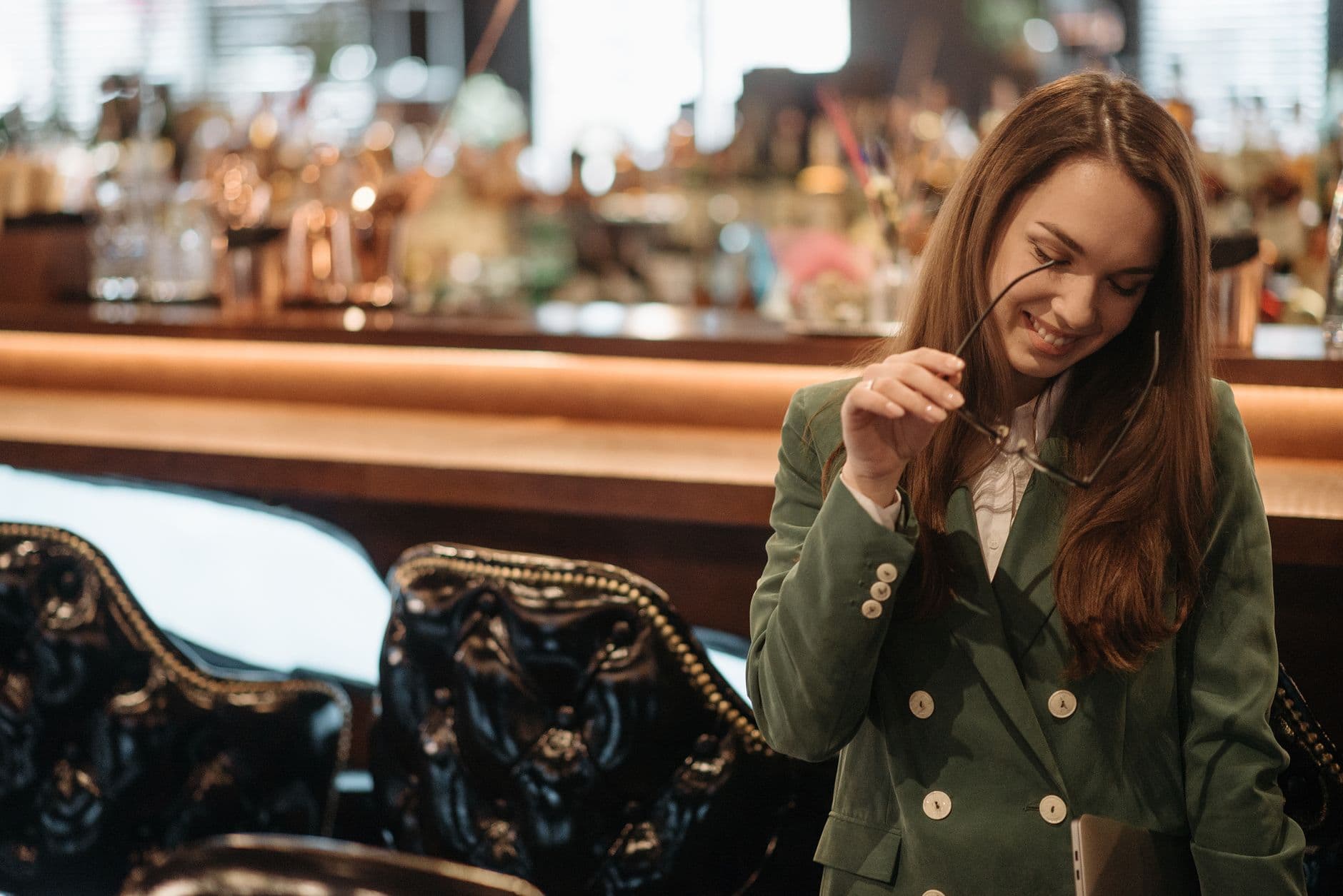 A smiling young woman in a green double-breasted blazer takes off her glasses in a warmly lit bar.