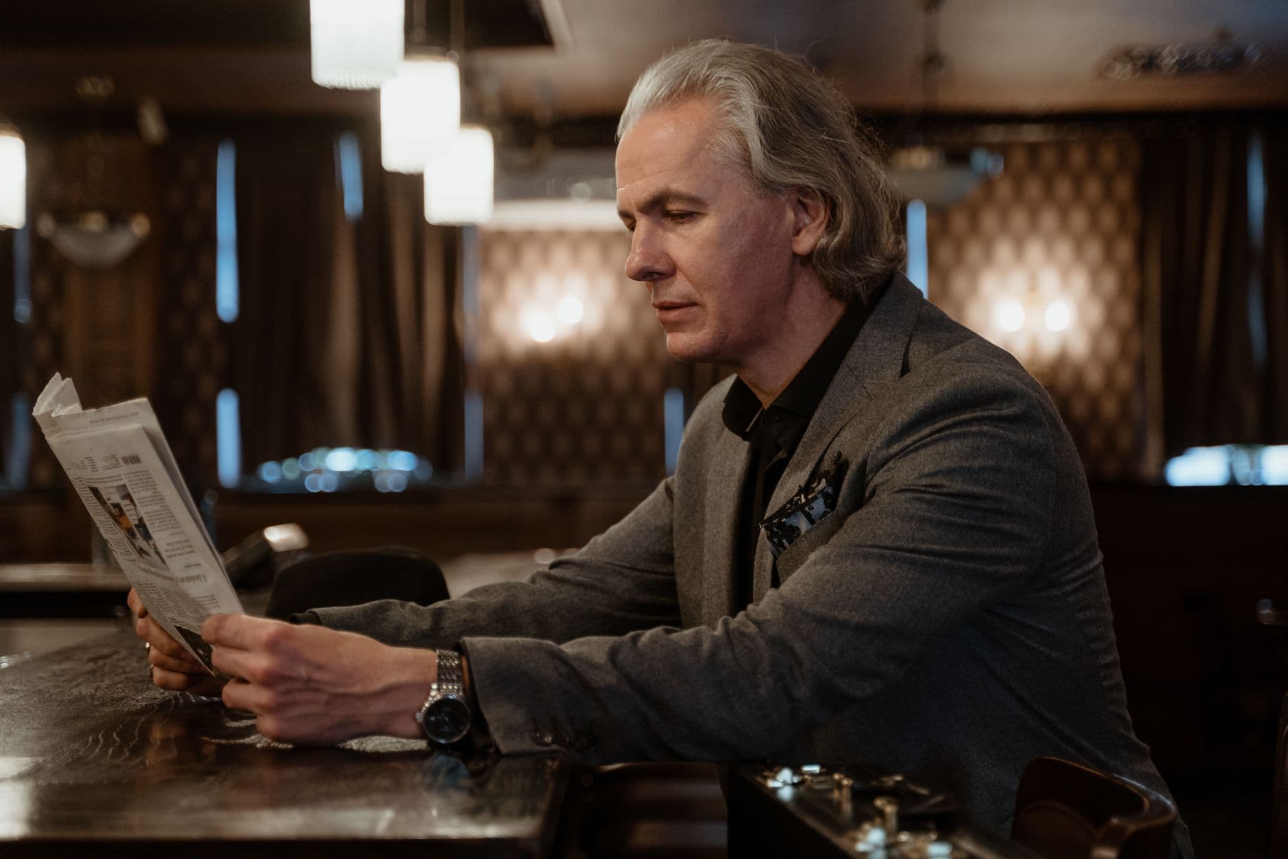 A silver-haired man in a grey jacket and wristwatch reads a newspaper at a dark wooden bar under soft lights.