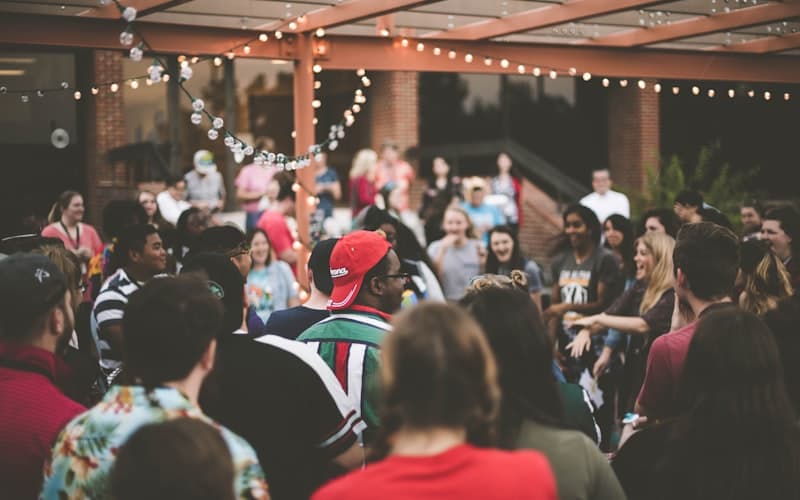 A crowd mingles under string lights at an outdoor party, one man in a red cap.