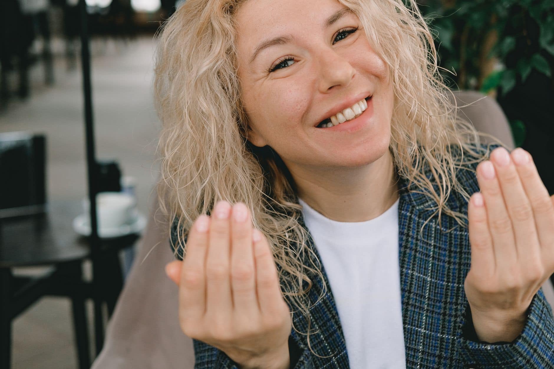 Een glimlachende vrouw met blonde krullen houdt beide handen voor zich bij een café.