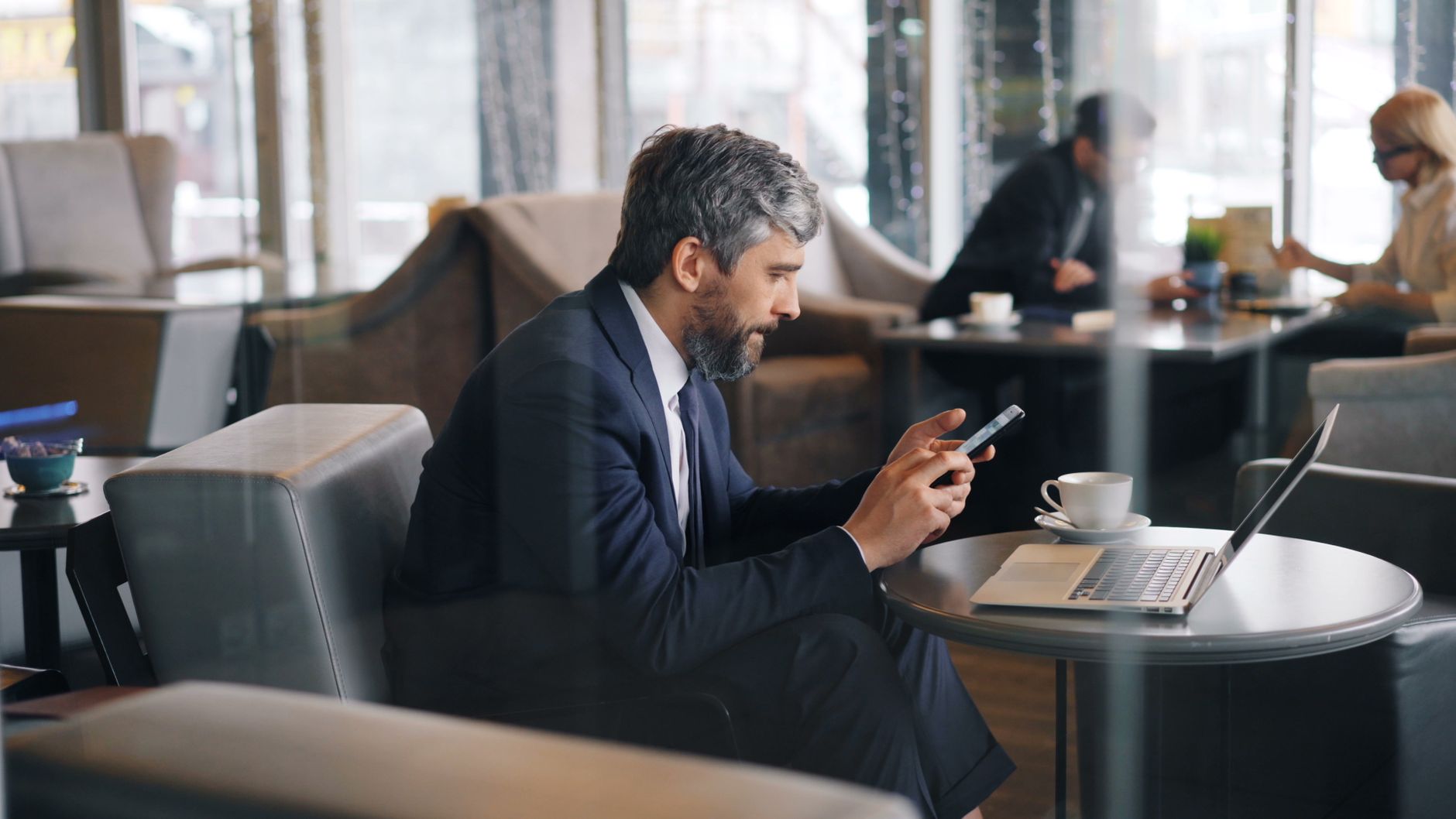 Elegant gråhårig man i mörkblå kostym sitter avslappnat i en stilfull kafélounge och läser ett meddelande på sin smartphone, med en kaffekopp på bordet
