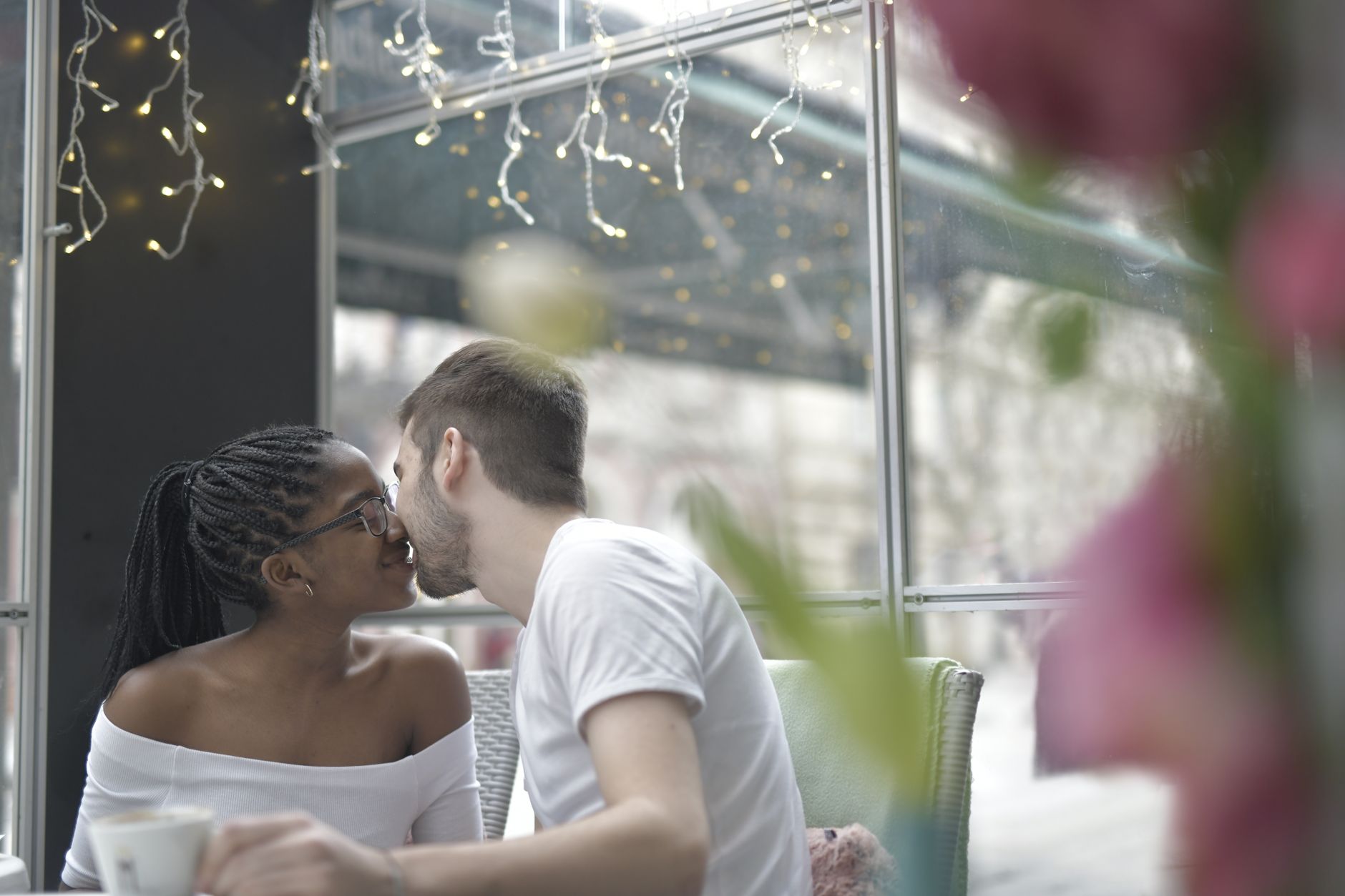 A young couple leans in for a kiss over coffee at a café window table strung with fairy lights, with the grey winter street visible through the glass behind them