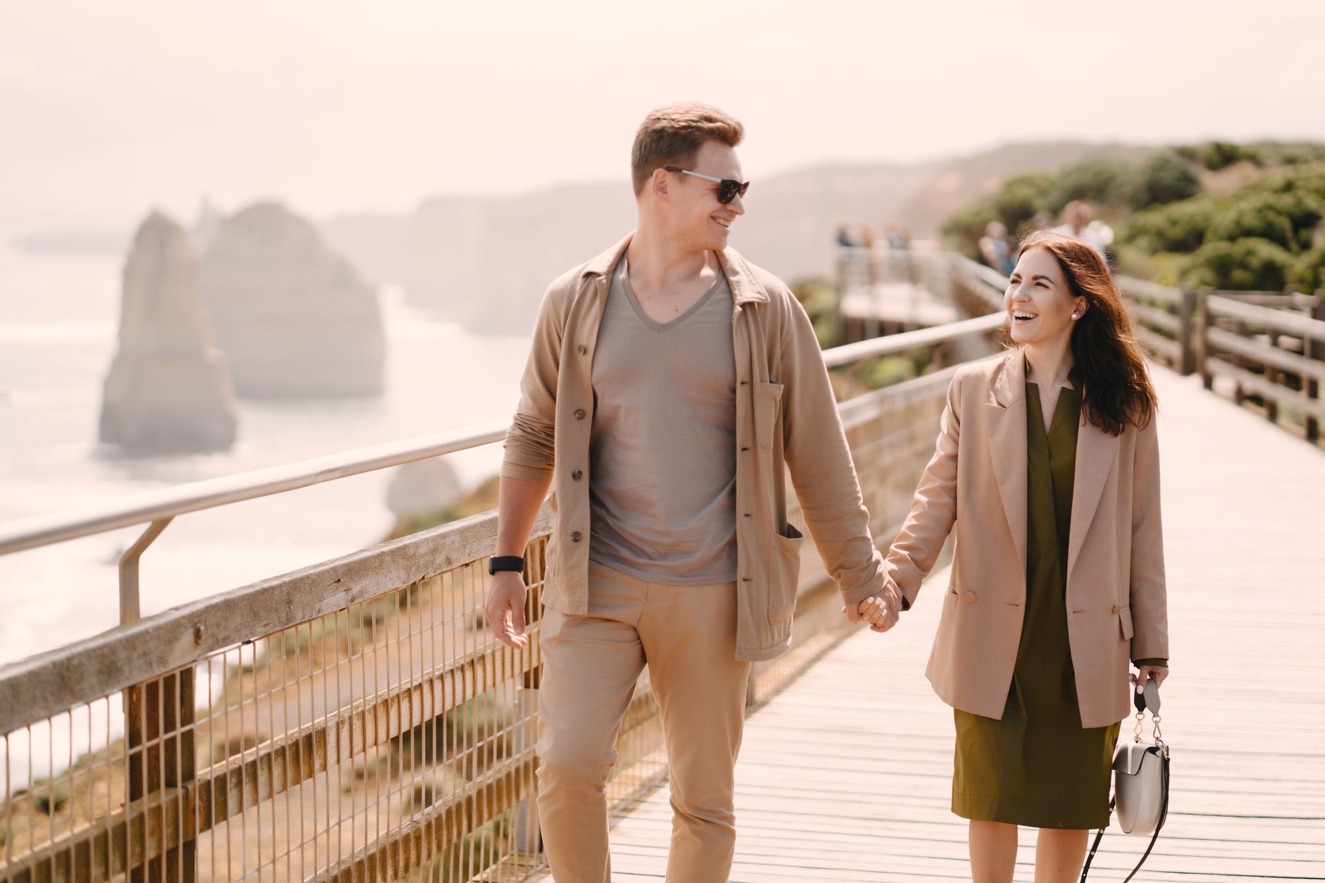 A laughing, stylishly dressed couple walks hand in hand along a wooden coastal boardwalk, with pale sea cliffs and the open sea hazy in the background