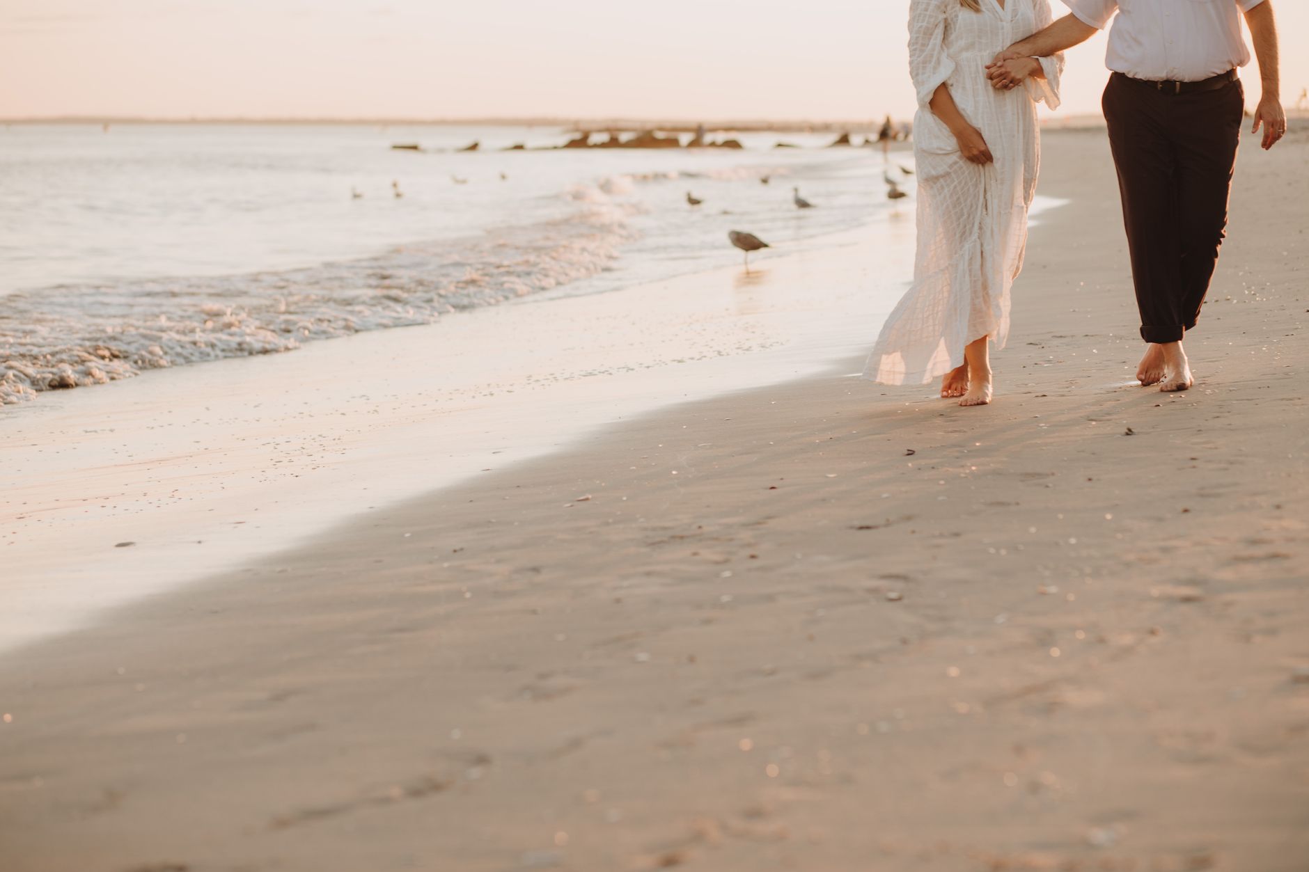 An elegant couple walks hand in hand along the wet sand at golden hour, her long white dress catching the breeze as seagulls gather at the water's edge.