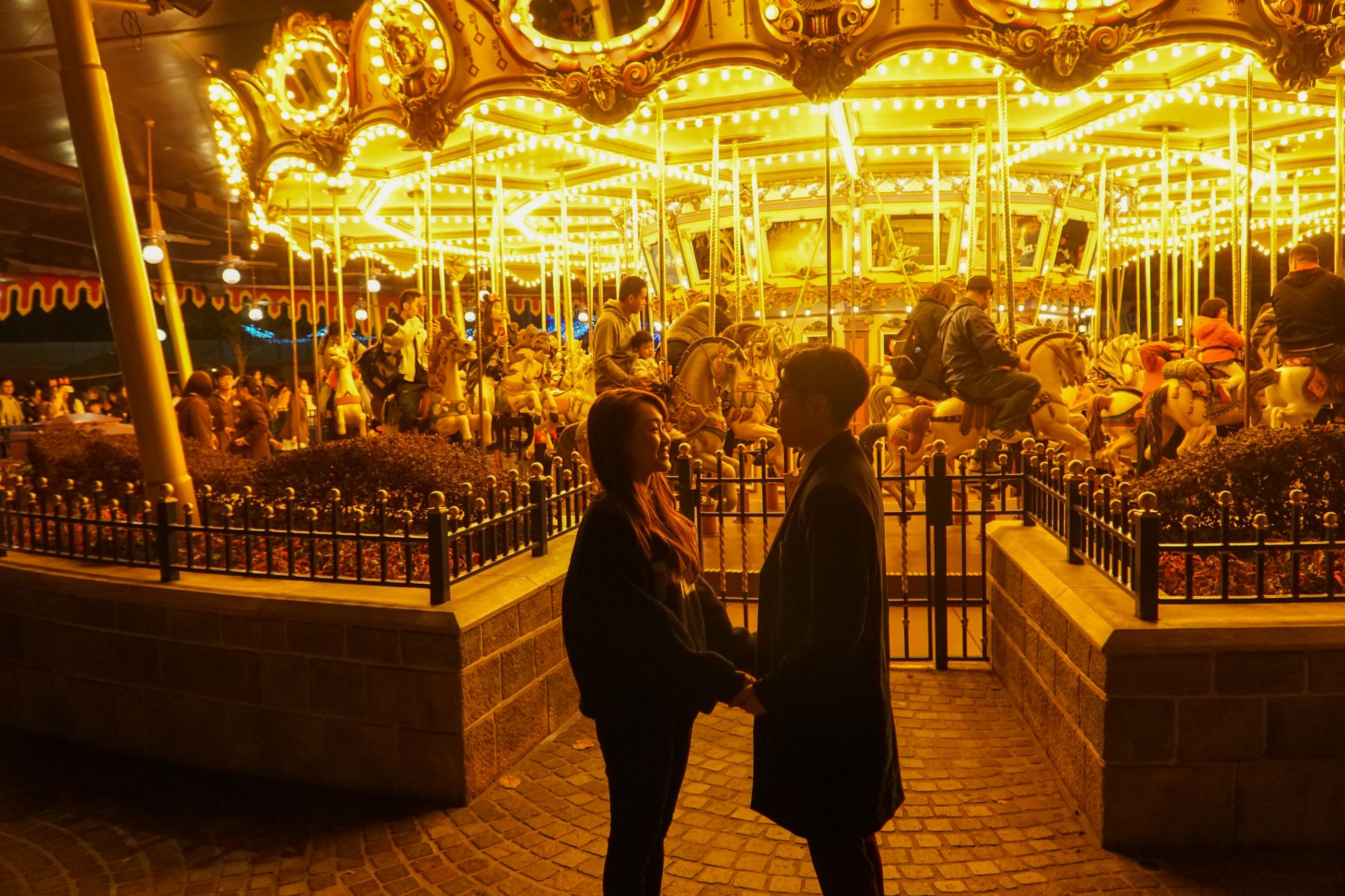Well-dressed couple holding hands and smiling at each other in front of a glowing carousel at an evening fair, bathed in warm golden light