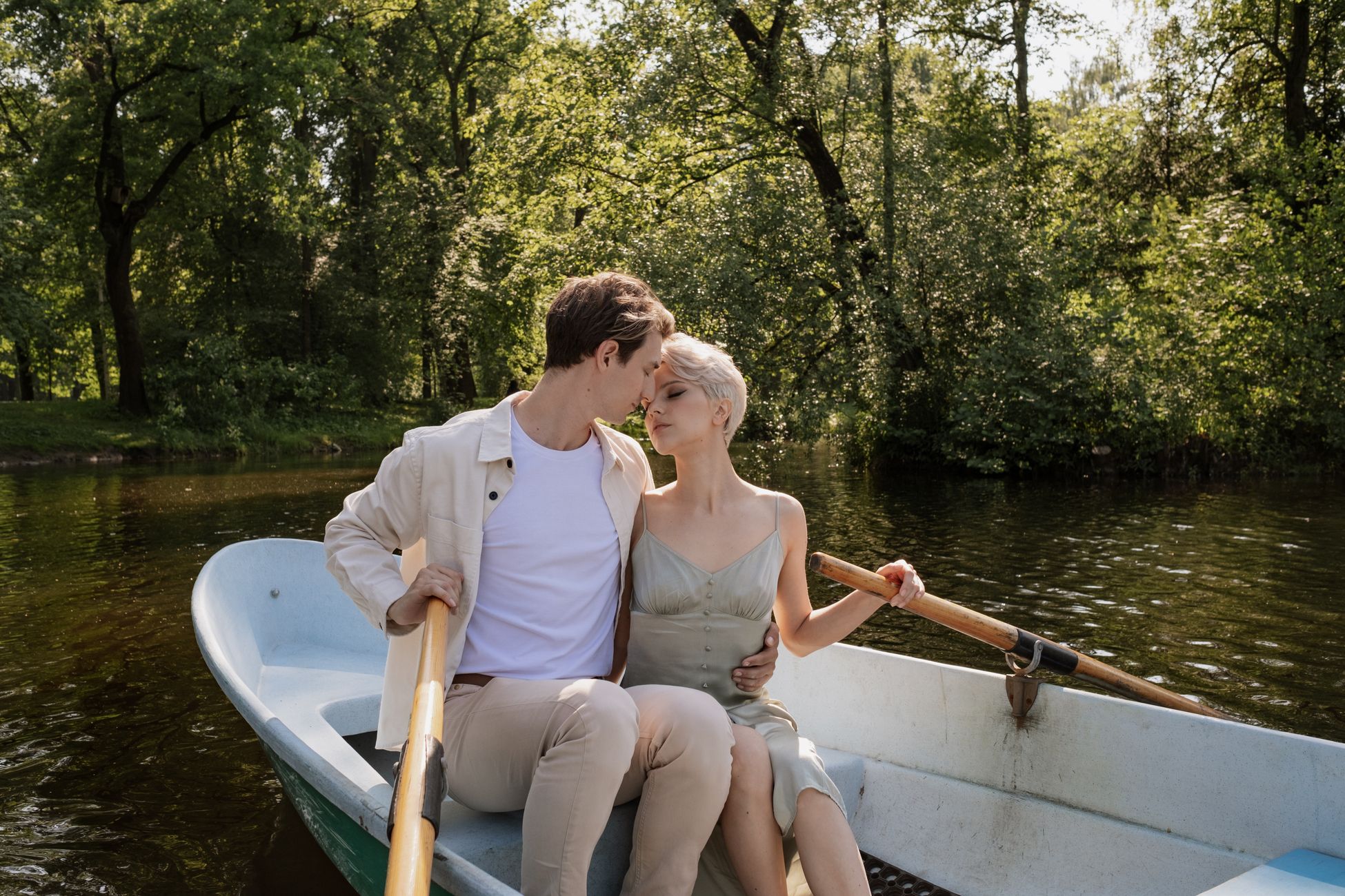 Stylish young couple sharing a tender moment in a rowboat on a calm tree-lined lake, each resting a hand on an oar in the summer sunlight