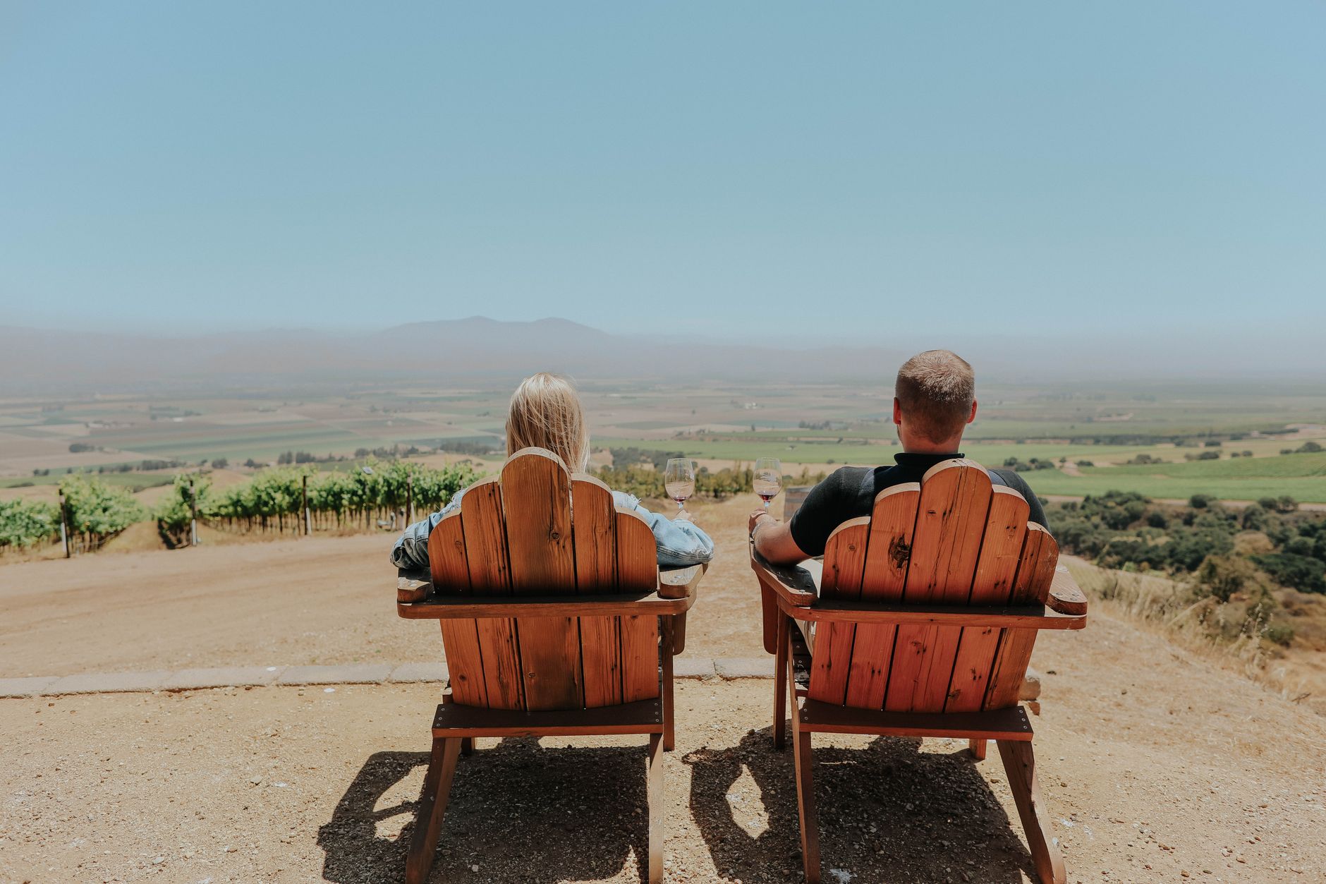 A couple sits side by side in wooden lookout chairs with glasses of wine, gazing out over green vineyard rows and a hazy summer valley