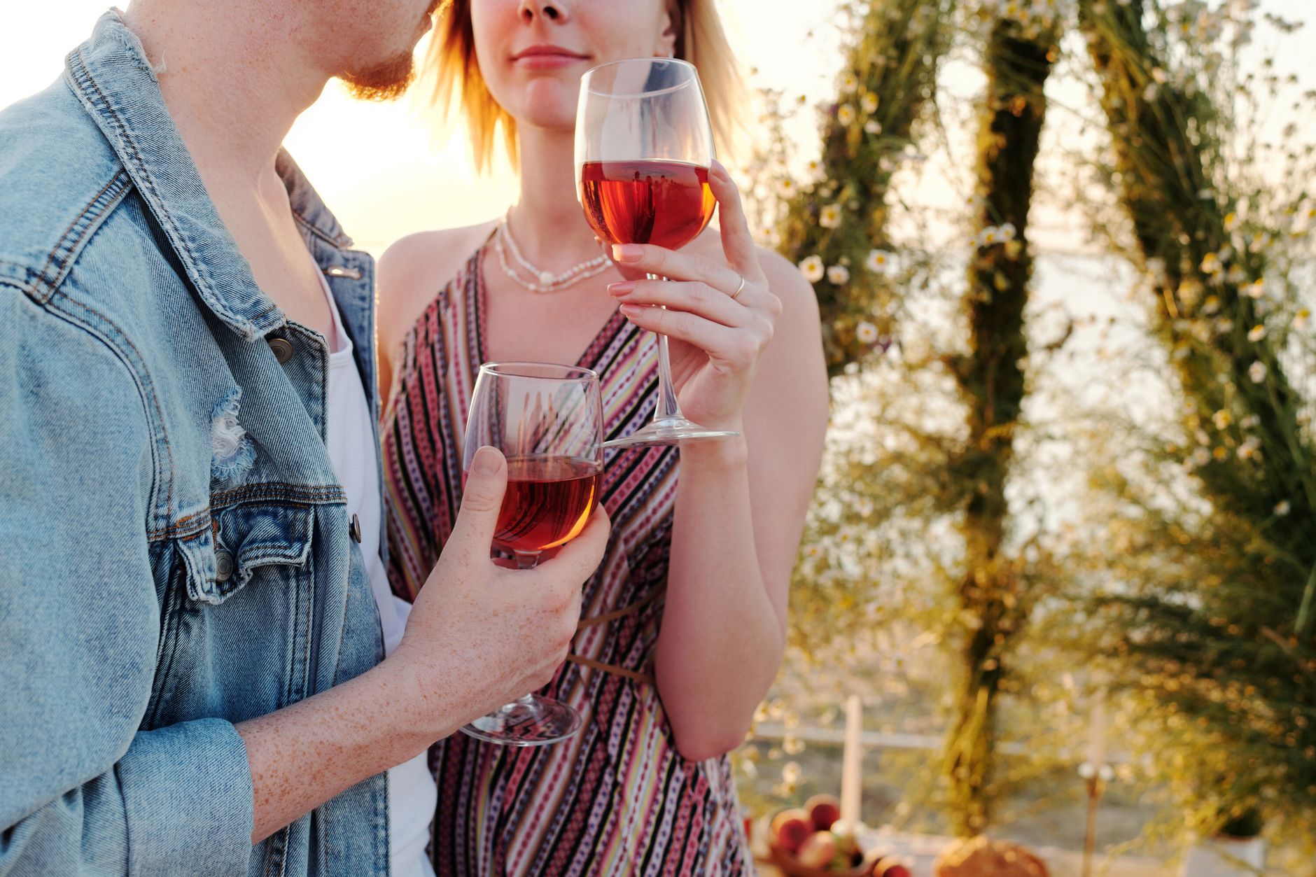 A young couple in summer clothes stands close together at golden hour, holding two glasses of rosé in front of a flower-covered arch