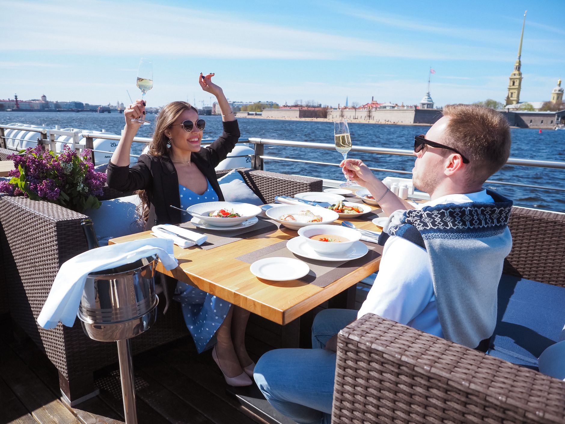Velklædt par skåler i champagne over et veldækket bord på en restaurantterrasse ved vandet — kvinden i lyseblå sommerkjole og mørk blazer, manden i lys skjorte med sweater over skuldrene, og havneudsigt i baggrunden